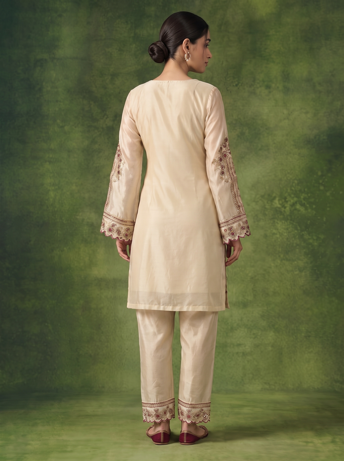 A woman shown from the back wearing a cream-colored traditional kurta and pant set with beige floral embroidery on the sleeves and hems, set against a textured green studio backdrop.