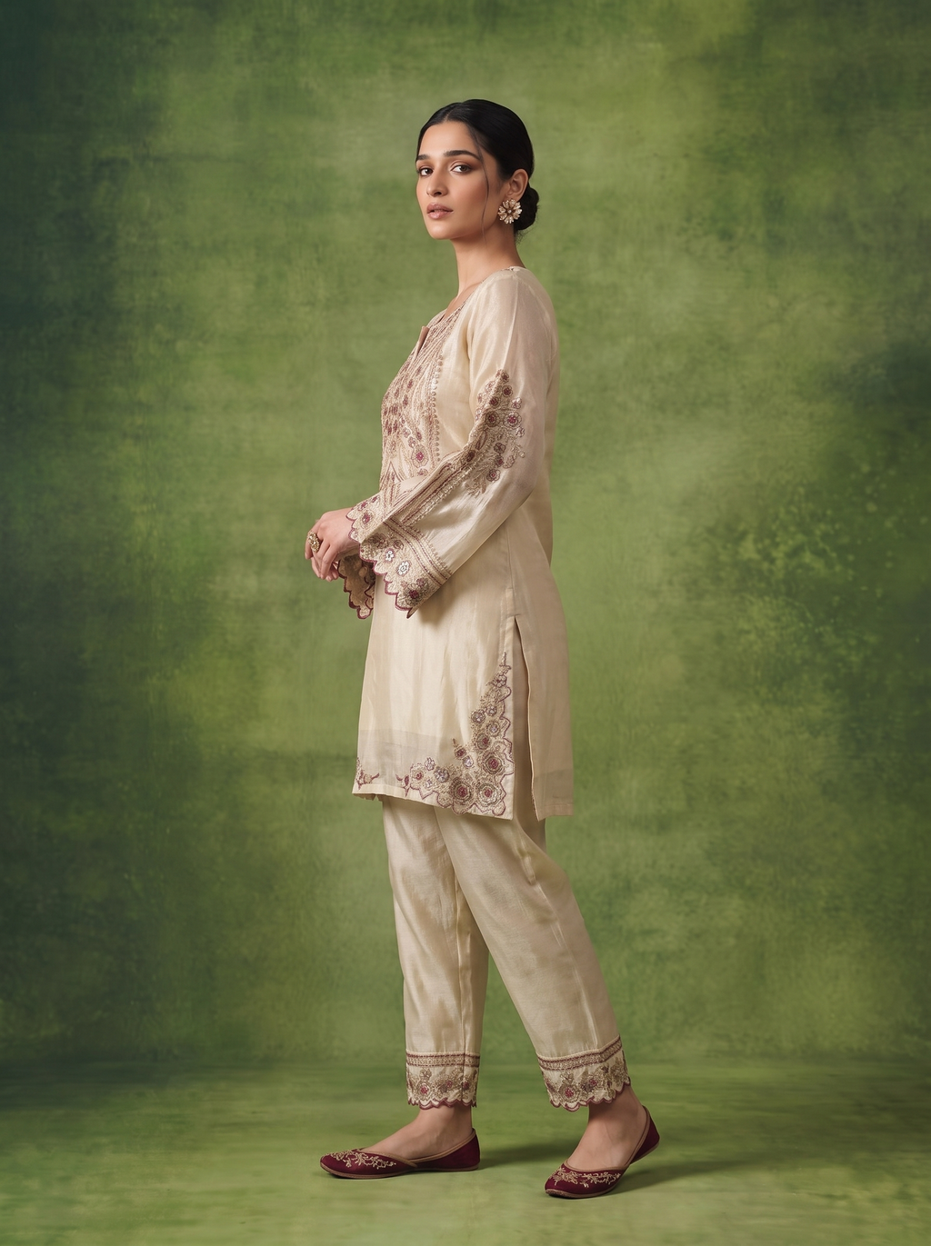 A woman modeling a traditional beige kurta and trouser set with floral embroidery, posed against a textured green studio backdrop.