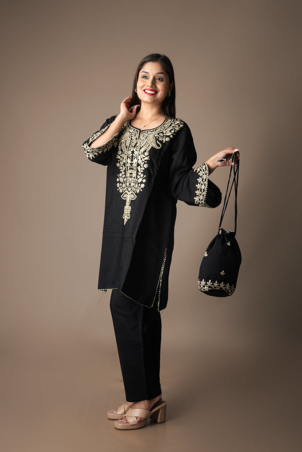 Woman wearing a black embroidered outfit holding a matching bag against a brown background