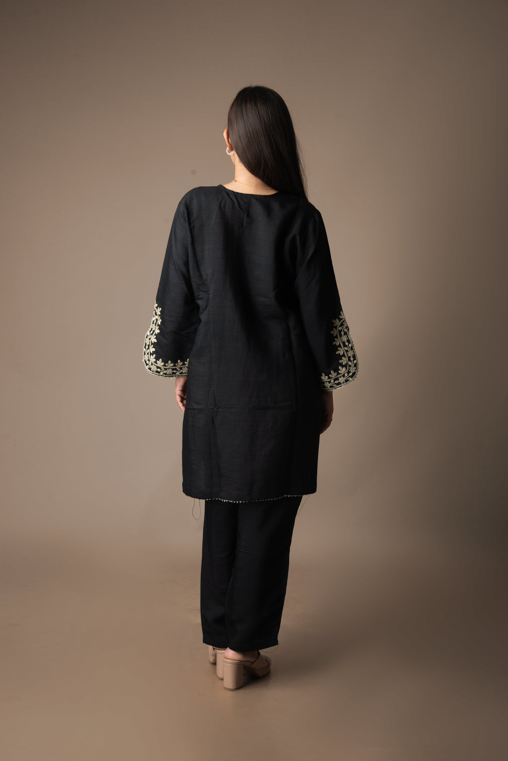 Person wearing a black outfit with floral embroidery on sleeves against a brown background
